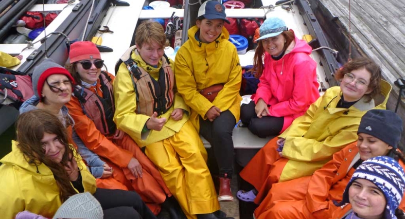 A group of students in life jackets and rain gear smile while sitting inside a sail boat. 